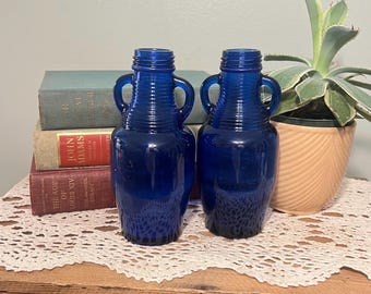 Peut inclure: Deux vases en verre bleu cobalt avec des anses. Les vases ont une texture côtelée et une forme d'amphore classique. Ils sont exposés sur une surface en bois avec des livres et une plante en pot en arrière-plan. Les vases mesurent environ 20 cm de haut.