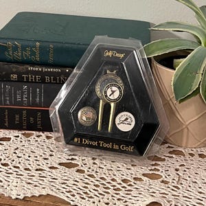 May include: A black and gold golf divot tool set in a clear plastic package. The set includes a divot tool and two ball markers. The text "Golf Design" and "#1 Divot Tool in Golf" are visible.