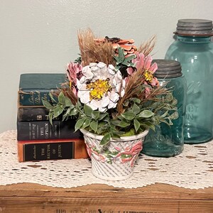 Può includere: Composizione floreale decorativa in un cestino bianco con nastri rosa, con fiori bianchi e gialli, accenti rosa e fogliame verde. Su una superficie di legno, con libri d'epoca e barattoli di vetro blu sullo sfondo.