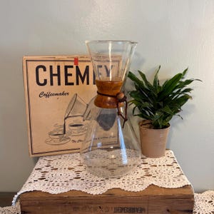 May include: A clear glass Chemex coffeemaker with a wooden collar and a leather tie, positioned in front of its original cardboard box featuring the text Chemex Coffeemaker and an illustration of coffee being poured into a cup.