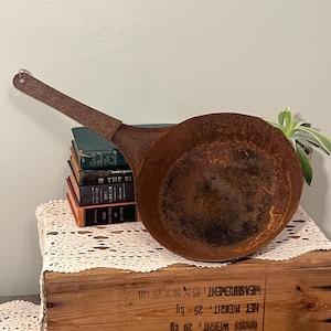 May include: An antique, rusty metal pan with a long handle. The pan is round with a deep bowl and shows signs of age and wear. The handle has a hole at the end for hanging. The pan is sitting on a wooden crate with books.