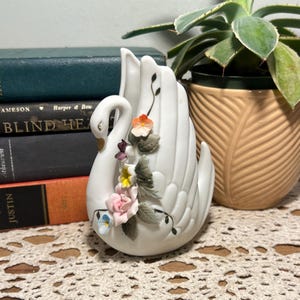 May include: A white ceramic swan figurine with a long neck and outstretched wings. The swan is adorned with colorful floral accents, including pink, yellow, and blue flowers. The swan is placed on a lace doily.