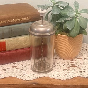 Puede incluir: Una coctelera de cristal vintage con tapa metálica y asa blanca. La coctelera tiene un cuerpo de cristal transparente y un agitador metálico en el interior. La coctelera está sobre una superficie de madera con libros y una planta en maceta al fondo.