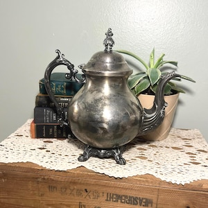 Puede incluir: Una tetera plateada vintage con asa y boquilla ornamentadas, apoyada sobre tres pies decorativos. La tetera tiene una tapa abovedada con un remate. El fondo incluye una pila de libros y una planta en maceta, todo sobre una superficie de madera.