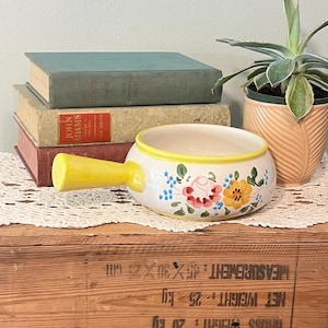 Puede incluir: Un cuenco de sopa de cerámica blanca con asa y borde amarillos, decorado con coloridos estampados florales. El cuenco está sobre un tapete de encaje blanco, con libros antiguos y una planta en maceta en el fondo.