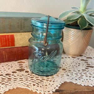 Op de afbeelding: Een vintage blauwe Ball Ideal pot met een draad handvat en deksel. De pot heeft het woord "Ball" in reliëf op de voorkant. De pot staat op een kanten kleedje, met boeken en een vetplant in een pot op de achtergrond.
