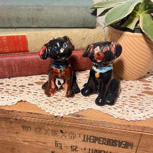 Puede incluir: Dos figuras de perro de cerámica vintage. Un perro es negro y marrón con un collar azul y una mancha blanca y negra en el pecho. El otro perro es negro con un collar azul y un dije dorado. Ambos están sobre un mantel de encaje blanco.