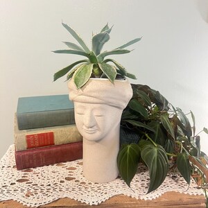 May include: A beige ceramic planter shaped like a serene face, with a turban-like head covering. The planter holds a green succulent plant. The planter is on a wooden surface, with vintage books and a lace doily in the background.
