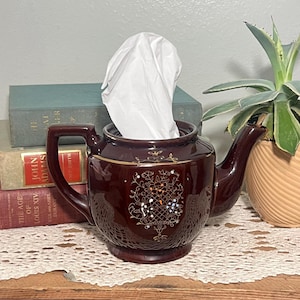 May include: A dark brown ceramic teapot-shaped tissue box holder with a handle and spout. The holder has gold trim and a floral design on the front. White tissues are visible inside the opening. A decorative item for the home.