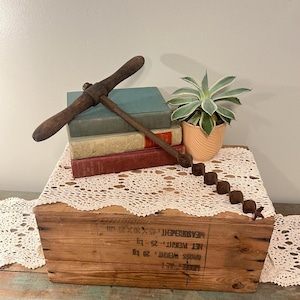 May include: A vintage wooden pickaxe rests atop a stack of antique books, with a small succulent in a peach pot. A string of small, dark brown decorative objects are arranged on a white lace doily, which sits on a wooden crate.