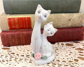 Puede incluir: Una figura de gato de cerámica blanca con dos gatos abrazándose. Los gatos tienen detalles faciales negros y naranjas y están sentados sobre una base con una rosa rosa y hojas verdes. La figura está frente a una pila de libros antiguos.