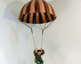 May include: A decorative hanging sculpture of a clown suspended from a brown and tan striped parachute. The clown figure is wearing a green outfit, a red hat, and has painted facial features. The parachute is attached by thin wires.