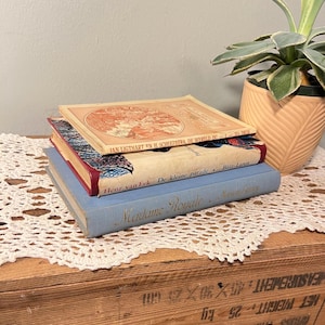 May include: A stack of three vintage books with different colored covers. The top book has a peach-colored cover with text. The middle book has a cream-colored cover with red trim. The bottom book is light blue with gold lettering.