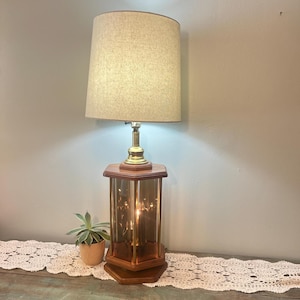 May include: A table lamp featuring a beige fabric drum shade, a brass-toned neck, and a hexagonal wooden base with amber-tinted glass panels. The lamp emits a warm glow from within the glass structure, illuminating the surrounding area.