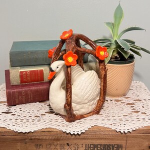 Könnte beinhalten: Eine weiße Keramik-Schwanfigur mit orangefarbenem Schnabel, umgeben von einem braunen, astähnlichen Rahmen, der mit orangefarbenen Blüten verziert ist. Die Figur steht auf einer Holzoberfläche, mit Büchern und einer Topfpflanze im Hintergrund.