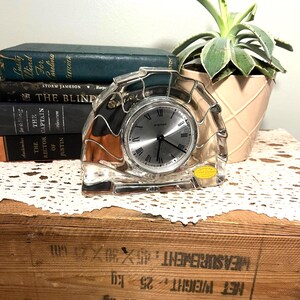 Puede incluir: Un reloj de cuarzo de cristal transparente con esfera plateada y números romanos. El reloj se exhibe sobre una caja de madera con libros y una suculenta en maceta en el fondo. El reloj tiene una pegatina "Genuine Lead Crystal".