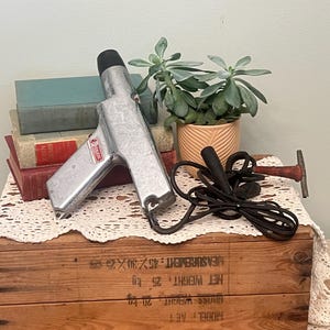 Puede incluir: Una pistola de pegamento plateada y negra vintage con un cable negro, apoyada en una caja de madera con libros y una suculenta en maceta. La pistola de pegamento tiene una etiqueta roja.