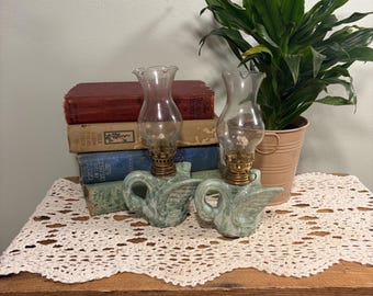 May include: Two matching oil lamps featuring light green ceramic swan-shaped bases with brass burners and clear glass chimneys. The lamps are positioned in front of a stack of four hardcover books and a potted plant in a light pink metal bucket.