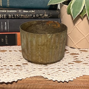 Puede incluir: Un recipiente cilíndrico de color latón envejecido con detalles acanalados verticales. El recipiente se encuentra sobre un mantel individual de encaje blanco, con libros en el fondo. El recipiente está vacío.