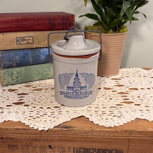 May include: A light gray ceramic canister with a matching lid and a dark metal wire handle. The front features a blue graphic illustration of Independence Hall with the text 1776 and 1976 on either side of the building name at the base.