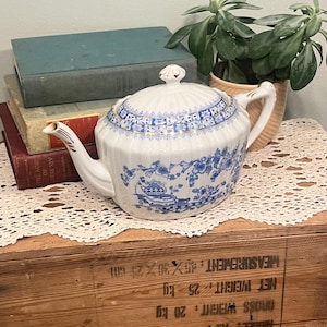Può includere: Teiera in porcellana bianca con un motivo floreale blu e rifiniture dorate. La teiera ha un coperchio con un pomello decorativo e un beccuccio curvo. È appoggiata su una cassa di legno.