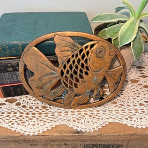 May include: An antique copper-toned metal fish-shaped trivet. The trivet features an oval frame with a detailed fish design in the center. The fish has textured scales and fins. The trivet is placed on a white lace doily.