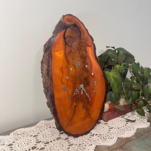 Puede incluir: Un reloj rústico de madera con borde natural. La esfera del reloj es de color marrón anaranjado cálido con números romanos y agujas plateadas. El reloj mide aproximadamente 30 cm de alto.