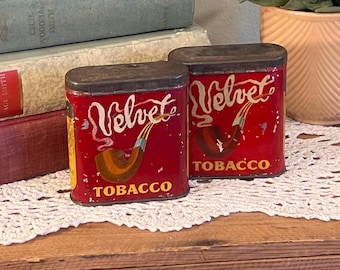 May include: Two vintage red tobacco tins with the word "Velvet" in white script and a brown pipe illustration. The word "TOBACCO" is printed in gold. The tins have a rectangular shape with a dark gray lid.