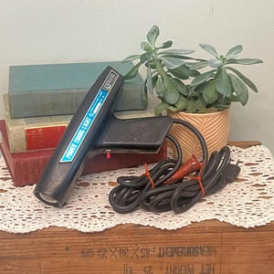 Puede incluir: Una herramienta negra Power Timing Light con un botón rojo y un cable negro largo. La herramienta tiene una etiqueta azul y blanca que dice "POWER TIMING LIGHT". Una planta suculenta en una maceta beige está detrás de la herramienta.