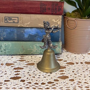 Könnte beinhalten: Eine Metallglocke mit einer silberfarbenen Figur eines Trompete spielenden Hofnarren, die auf einem messingfarbenen Glockensockel montiert ist. Der Narr trägt eine spitze Mütze und detaillierte Kleidung, die Glocke hat eine glatte Öffnung.