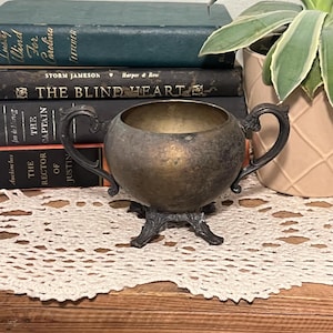 Puede incluir: Azucarero antiguo de color plateado con dos asas ornamentadas y una base decorativa. El cuenco está sobre un tapete de encaje blanco, con una pila de libros antiguos en el fondo. Los libros son de varios colores, incluyendo azul y negro.