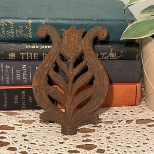 May include: A vintage, ornate, cast iron decorative piece with a floral design. The brown, weathered metalwork features a central stem with leaf details and a tulip-shaped top. The piece is set against a backdrop of antique books.
