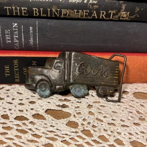 以下が含まれることがあります： 配送トラックの形をしたヴィンテージの金属製ベルトバックル。トラックはブロンズ色で、側面に「Coors」の文字が入っています。車輪は青緑色の緑青があります。バックルは古い本の背景に配置されています。