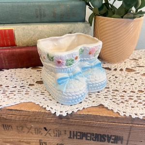 May include: A light blue ceramic baby shoe planter with a textured design. The planter is decorated with pink and white flowers, green leaves, and a light blue bow. The planter is sitting on a white lace doily.