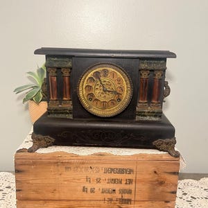 Puede incluir: Reloj de chimenea negro antiguo con esfera dorada y números romanos. El reloj presenta columnas decorativas y detalles ornamentados. El reloj está sobre una caja de madera.