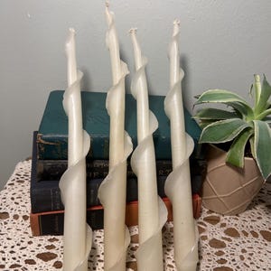 Puede incluir: Cuatro velas en espiral de color marfil. Cada vela presenta un diseño único en forma de cinta que se envuelve alrededor del cuerpo principal. Las velas se exhiben sobre una mesa con libros y una planta de fondo. Las velas miden unos 25 cm de alto.