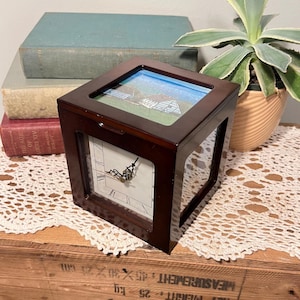 Puede incluir: Un reloj cuadrado de madera oscura con esfera blanca y manecillas negras. El reloj tiene una tapa de cristal con una pintura de un edificio y un cielo azul. El reloj está sobre una superficie de madera.