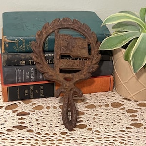 May include: A vintage, brown metal trivet with a flag design. The trivet has a circular frame with a decorative leaf pattern and a handle. It is resting on a white lace tablecloth, with a stack of old books in the background.