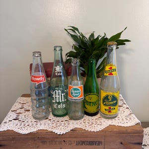 May include: Five vintage glass soda bottles. From left to right: a clear Howdy bottle, a green Mr. Cola bottle, a clear Green Spot bottle, a green Quench bottle, and a clear Vernors bottle with a yellow label reading 1 Calorie.
