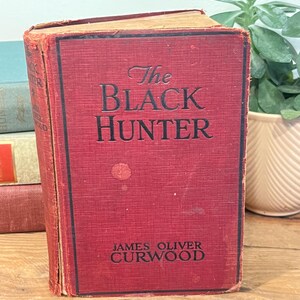 K&ouml;nnte beinhalten: Ein Vintage-Hardcover-Buch mit dem Titel "The Black Hunter" von James Oliver Curwood. Das Buch hat einen roten Einband mit schwarzer Schrift und weist Gebrauchsspuren auf. Das Buch wird auf einer Holzoberfl&auml;che pr&auml;sentiert.