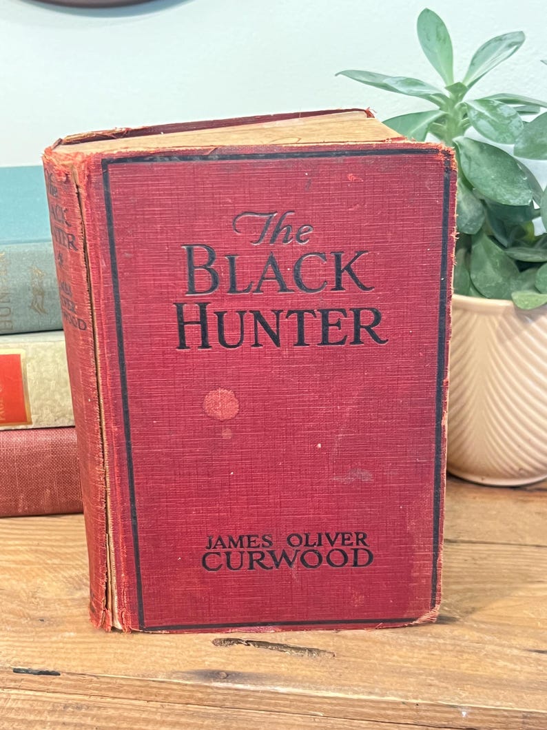 K&ouml;nnte beinhalten: Ein Vintage-Hardcover-Buch mit dem Titel "The Black Hunter" von James Oliver Curwood. Das Buch hat einen roten Einband mit schwarzer Schrift und weist Gebrauchsspuren auf. Das Buch wird auf einer Holzoberfl&auml;che pr&auml;sentiert.