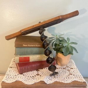 May include: An antique auger drill with a wooden handle and a dark metal spiral bit. The drill is positioned above a stack of vintage books, including one titled "Gem Hunter's Guide". A small potted succulent plant sits nearby.