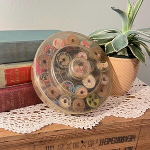 Könnte beinhalten: Eine transparente, kreisförmige Nähkiste mit Deckel, gefüllt mit bunten Garnrollen. Das Set steht auf einer Holzoberfläche mit einer weißen Spitzendecke. Bücher und eine Topfpflanze befinden sich im Hintergrund.