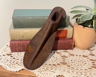 May include: A weathered, metal shoe last, showing signs of rust, with a long, pointed form and a rectangular aperture at the top. It rests on a wooden surface with a white lace doily, with antique books in the background.