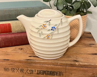 May include: Cream-coloured ceramic teapot with a ribbed design and a lid. The teapot features hand-painted floral accents in blue, orange, and green. It has a curved handle and a spout. The teapot is displayed on a wooden surface.