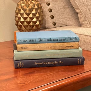 May include: A stack of four vintage books with titles including "The Gentleman from California" and "Pretend You Don't See Her." The books are in various colors, including blue, tan, and gray, and are stacked on a wooden surface.