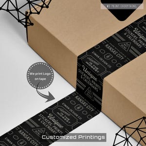 May include: A brown cardboard box with black tape wrapped around it. The tape features white text that reads "Unique Stickers, Prints & Calendars" and "Kansas City." The image also includes the text "Customized Printings."