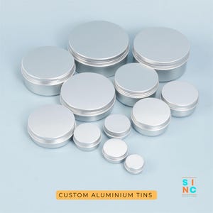 Op de afbeelding: Een verzameling zilveren aluminium blikken met deksels in verschillende maten. De blikken zijn gerangschikt op een lichtblauwe ondergrond. De tekst "CUSTOM ALUMINIUM TINS" staat in een oranje rechthoek onderaan de afbeelding.
