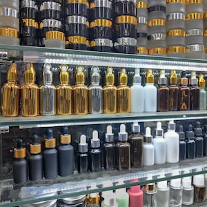 Op de afbeelding: Een display van diverse cosmetische containers, waaronder glazen flessen met druppelaars, potten met deksels en kleine metalen blikjes. De flessen zijn er in goud, zilver, zwart en helder glas, met gouden of zilveren doppen. De potten hebben zwarte, gouden en zilveren deksels.