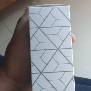 Puede incluir: Una caja rectangular blanca con un patr&oacute;n geom&eacute;trico gris. La caja est&aacute; sostenida por una persona. El fondo est&aacute; desenfocado.